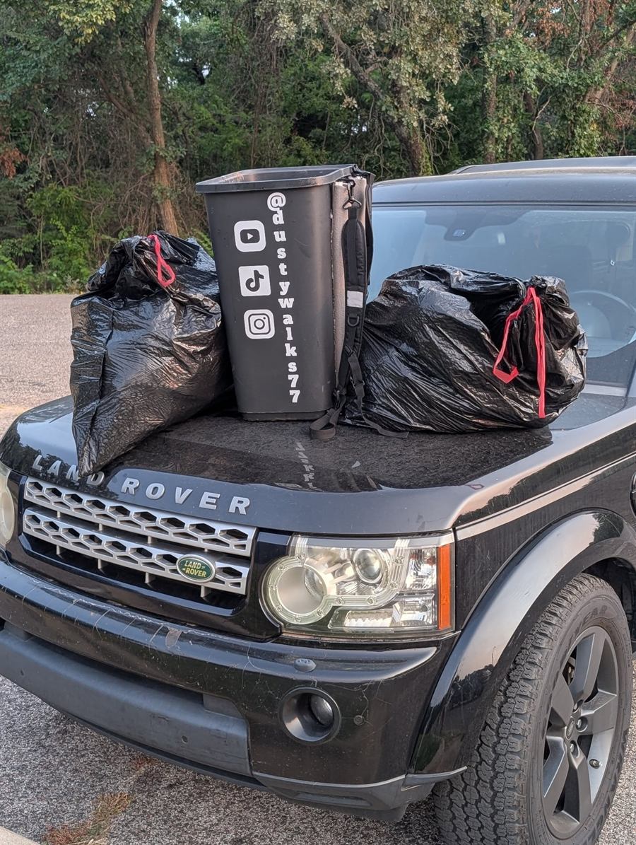Two full black trash bags either side of a bin marked @dustywalks77, sitting on the hood of a black Land Rover at the edge of a wooded road.