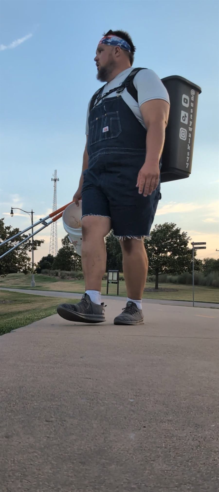 Dusty walking a paved park path at sunset in overalls and a stars-and-stripes bandana, a trash can strapped to his back marked @dustywalks77, a grabber and a bucket in his hand.